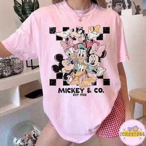 May include: A light pink t-shirt featuring a graphic of Mickey Mouse and friends, including Minnie Mouse, Donald Duck, and Goofy. The design is set against a black and white checkered background, with "Mickey & Co. EST 1928" printed below.