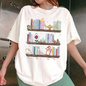 Puede incluir: Camiseta color crema con una ilustraci&oacute;n caprichosa de tres estanter&iacute;as. Cada estanter&iacute;a est&aacute; llena de libros coloridos y objetos tem&aacute;ticos, incluyendo una rana, una rosa, una l&aacute;mpara y un drag&oacute;n. El dise&ntilde;o tiene un tema de cuento de hadas.
