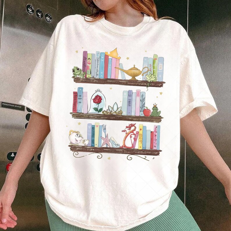 Puede incluir: Camiseta color crema con una ilustraci&oacute;n caprichosa de tres estanter&iacute;as. Cada estanter&iacute;a est&aacute; llena de libros coloridos y objetos tem&aacute;ticos, incluyendo una rana, una rosa, una l&aacute;mpara y un drag&oacute;n. El dise&ntilde;o tiene un tema de cuento de hadas.