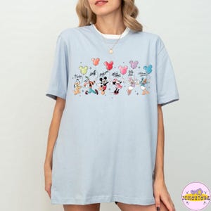以下が含まれることがあります： ミッキーマウスやミニーマウスなど、ディズニーキャラクターが風船を持っているカラフルなグラフィックが特徴のライトブルーのTシャツ。キャラクターの名前が上に書かれています。半袖でクルーネックです。