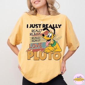 以下が含まれることがあります： ディズニーの漫画の犬、プルートが描かれたマスタードイエローのTシャツ。「I Just Really Really Really Really Love Pluto」の文字。プルートは黄色で、黒い耳と舌が出ている。テキストは黒とオレンジ。