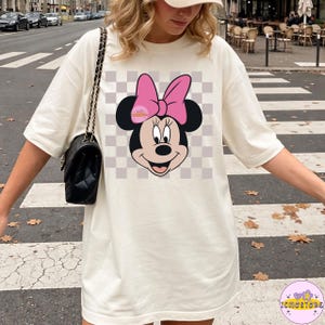 Puede incluir: Camiseta color crema con un gr&aacute;fico de Minnie Mouse. El dise&ntilde;o incluye la cara de Minnie con un lazo rosa, sobre un fondo a cuadros. La camiseta tiene mangas cortas y un corte holgado, adecuada para uso casual.