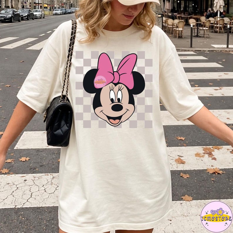 Puede incluir: Camiseta color crema con un gr&aacute;fico de Minnie Mouse. El dise&ntilde;o incluye la cara de Minnie con un lazo rosa, sobre un fondo a cuadros. La camiseta tiene mangas cortas y un corte holgado, adecuada para uso casual.