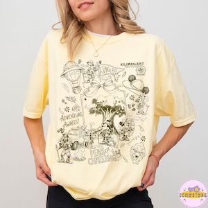 Puede incluir: Una camiseta de manga corta de color amarillo pálido con un estampado gráfico en marrón oscuro de personajes de dibujos animados con equipo de safari, un árbol grande, un vehículo y el texto Adventure Awaits, Jambo, Kilimanjaro Safaris y Peak Challenge.
