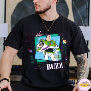 May include: Black t-shirt featuring a graphic of Buzz Lightyear from Toy Story. The design includes the word "BUZZ" and the word "Her" with a retro 90s style design. The character is in a square frame with a blue background.