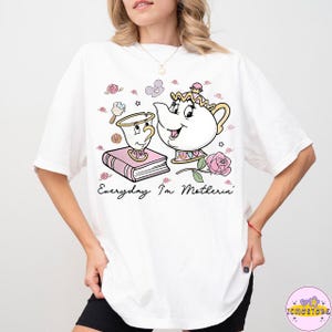 May include: White t-shirt featuring a cartoon illustration of a teapot and teacup with the text "Everyday I'm Motherin'". The design includes pink roses and a book. The shirt is a casual, comfortable style.