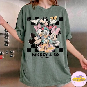 May include: Green t-shirt featuring a graphic of Mickey Mouse and friends. The design includes Mickey, Minnie, Donald, Daisy, Goofy, and Pluto. The text "MICKEY & CO. EST 1928" is printed below the characters. The shirt has a relaxed fit.