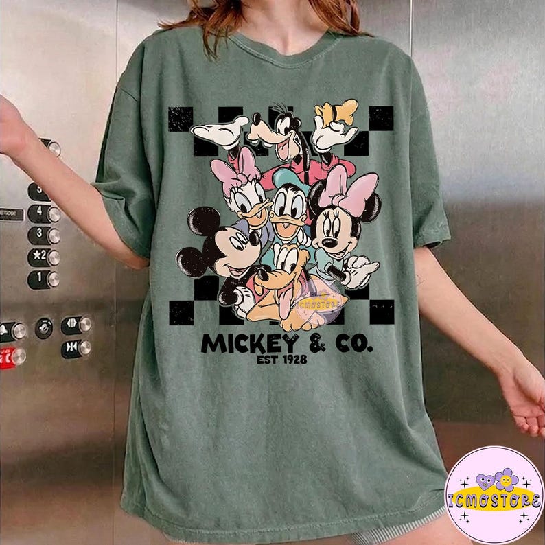 May include: Green t-shirt featuring a graphic of Mickey Mouse and friends. The design includes Mickey, Minnie, Donald, Daisy, Goofy, and Pluto. The text "MICKEY & CO. EST 1928" is printed below the characters. The shirt has a relaxed fit.