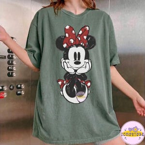 May include: Green t-shirt featuring a Minnie Mouse graphic. The graphic shows Minnie Mouse with a red bow with white polka dots. The t-shirt is a relaxed fit and has short sleeves. The t-shirt is a solid green color.
