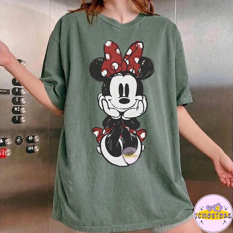 May include: Green t-shirt featuring a Minnie Mouse graphic. The graphic shows Minnie Mouse with a red bow with white polka dots. The t-shirt is a relaxed fit and has short sleeves. The t-shirt is a solid green color.