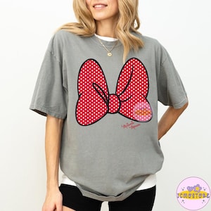 May include: A gray t-shirt featuring a large red polka-dot bow graphic with a black outline. The bow has a smaller pink graphic on the right side and the words "Minnie Mouse" in cursive below the bow.