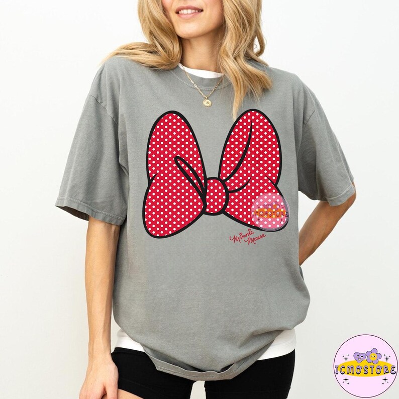May include: A gray t-shirt featuring a large red polka-dot bow graphic with a black outline. The bow has a smaller pink graphic on the right side and the words "Minnie Mouse" in cursive below the bow.