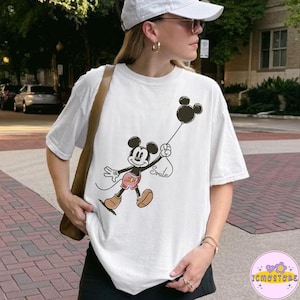 Könnte beinhalten: Weißes T-Shirt mit einem Cartoon-Mickey-Mouse-Design, das einen schwarzen Ballon hält. Auf der Schnur des Ballons steht das Wort "Smile". Mickey trägt rote Shorts und braune Schuhe. Das T-Shirt ist ein lässiger Stil.