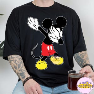 Puede incluir: Camiseta negra con un dise&ntilde;o de Mickey Mouse haciendo el dab. Mickey est&aacute; representado en sus colores cl&aacute;sicos: negro, rojo, amarillo y blanco. La camiseta tiene un corte holgado y mangas cortas. El dise&ntilde;o incluye el texto "ICMOSTORE" en el zapato.