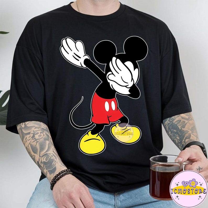 Puede incluir: Camiseta negra con un dise&ntilde;o de Mickey Mouse haciendo el dab. Mickey est&aacute; representado en sus colores cl&aacute;sicos: negro, rojo, amarillo y blanco. La camiseta tiene un corte holgado y mangas cortas. El dise&ntilde;o incluye el texto "ICMOSTORE" en el zapato.