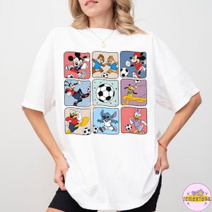 May include: White t-shirt featuring a grid of colorful squares with Disney characters playing soccer. Mickey Mouse, Chip and Dale, Goofy, Donald Duck, Stitch, and Daisy Duck are depicted with soccer balls in a playful design.