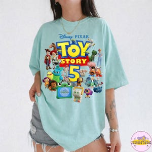 May include: A light blue t-shirt featuring the Disney Pixar Toy Story 5 logo and various character illustrations. The shirt has a crew neck and short sleeves. The design includes characters like Woody, Buzz Lightyear, and others from the animated film series.