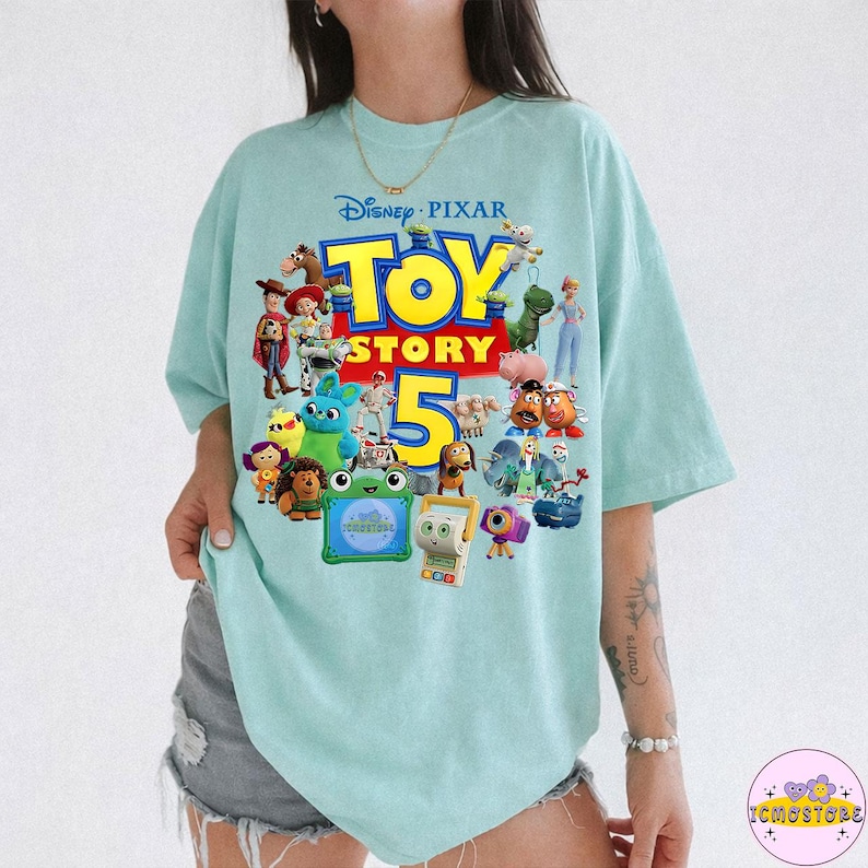 May include: A light blue t-shirt featuring the Disney Pixar Toy Story 5 logo and various character illustrations. The shirt has a crew neck and short sleeves. The design includes characters like Woody, Buzz Lightyear, and others from the animated film series.