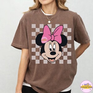 Puede incluir: Camiseta marr&oacute;n con un gr&aacute;fico de Minnie Mouse, lazo rosa y fondo a cuadros. La camiseta tiene mangas cortas y cuello redondo. El gr&aacute;fico est&aacute; centrado en la parte delantera.