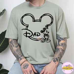 May include: Sage green t-shirt featuring a black outline of Mickey Mouse ears with the word "Dad" in a Disney-inspired font. A seated Mickey Mouse graphic is on the right. The shirt is a casual, everyday wear item.