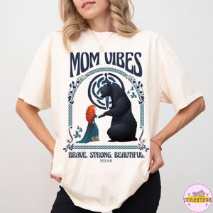 Op de afbeelding: Crèmekleurig T-shirt met een afbeelding van een beer en een meisje met rood haar, met de woorden "MOM VIBES" erboven en "BRAVE. STRONG. BEAUTIFUL." eronder. Het shirt heeft een ronde hals en korte mouwen.