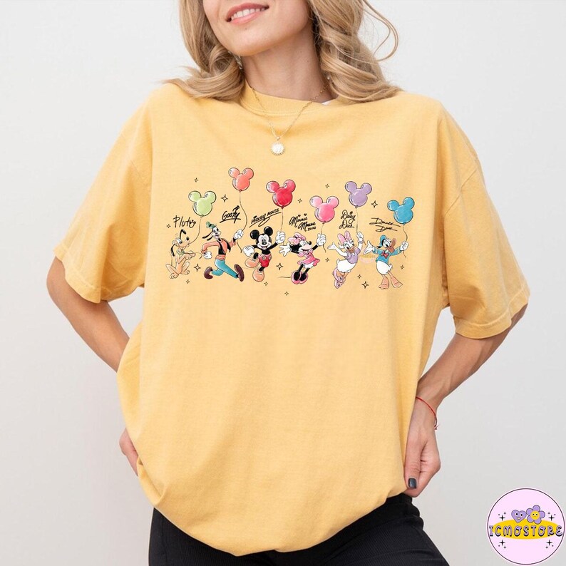 以下が含まれることがあります： マスタードイエローのTシャツ。ディズニーのキャラクターが風船のブーケを持っているグラフィックが特徴です。キャラクターにはプルート、グーフィー、ミッキーマウス、ミニーマウス、デイジーダック、ドナルドダックが含まれます。風船は様々な色です。