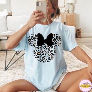 May include: Light blue t-shirt featuring a Minnie Mouse design. The design includes a black bow and a leopard print pattern in shades of brown, black, and white. The shirt is a casual, short-sleeved style.