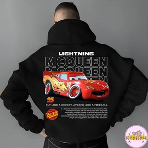 May include: Black hoodie featuring Lightning McQueen from the Cars movie franchise. The design includes the text "LIGHTNING MCQUEEN" and a graphic of the red race car with the number 95. The hoodie also has the Rust-eze logo and the phrase "FLY LIKE A ROCKET, ATTACK LIKE A FIREBALL."