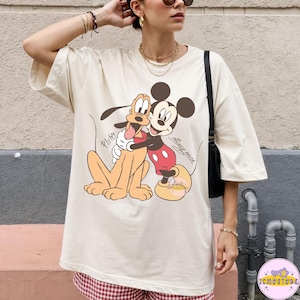 May include: Cream-colored oversized t-shirt featuring a graphic of Mickey Mouse hugging Pluto. Mickey Mouse is in his classic red shorts and yellow shoes. Pluto is a light brown color. The words "Pluto" and "Mickey Mouse" are also on the shirt.
