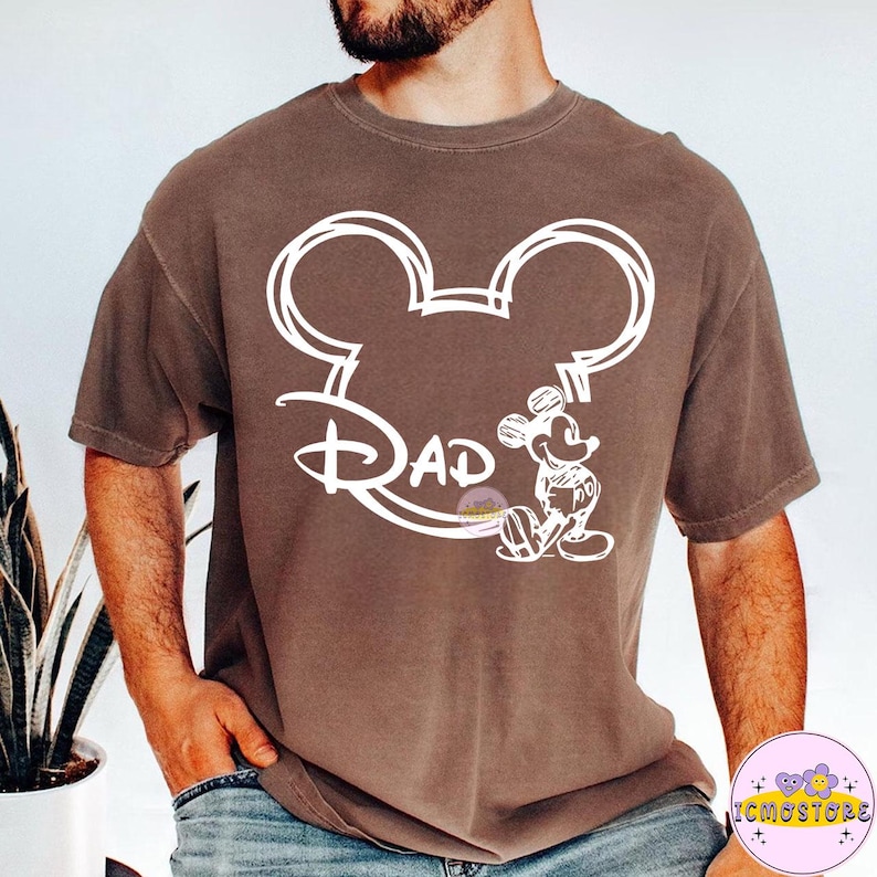 May include: A brown t-shirt featuring a white outline of Mickey Mouse ears with the word "Dad" in a Disney-inspired font. A small illustration of Mickey Mouse is on the right side of the design. The shirt has short sleeves.