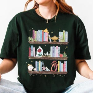 Puede incluir: Camiseta verde oscuro con una ilustraci&oacute;n caprichosa de tres estanter&iacute;as de madera llenas de libros coloridos y objetos tem&aacute;ticos de cuentos de hadas. El dise&ntilde;o incluye una rana, una rosa en un vaso, una l&aacute;mpara y un drag&oacute;n.