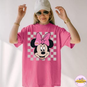 Puede incluir: Camiseta rosa con un dise&ntilde;o de Minnie Mouse. El dise&ntilde;o incluye la cara de Minnie, un lazo rosa y un fondo a cuadros. La camiseta es de color rosa liso y tiene mangas cortas.