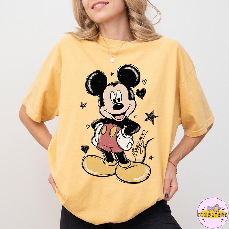 May include: Yellow t-shirt featuring a Mickey Mouse graphic. The cartoon character is drawn in black and white with red shorts and yellow shoes. The shirt has a relaxed fit and is adorned with hearts and stars.