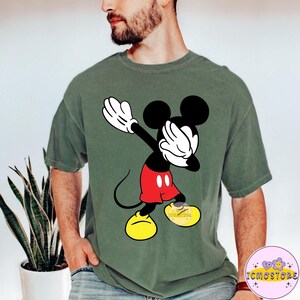Puede incluir: Camiseta verde oliva con un gr&aacute;fico de Mickey Mouse haciendo dab. Mickey se muestra en negro, blanco, rojo y amarillo. La camiseta tiene un corte holgado y un lavado vintage. El gr&aacute;fico es una versi&oacute;n l&uacute;dica del personaje cl&aacute;sico.
