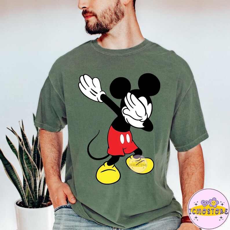 Puede incluir: Camiseta verde oliva con un gr&aacute;fico de Mickey Mouse haciendo dab. Mickey se muestra en negro, blanco, rojo y amarillo. La camiseta tiene un corte holgado y un lavado vintage. El gr&aacute;fico es una versi&oacute;n l&uacute;dica del personaje cl&aacute;sico.