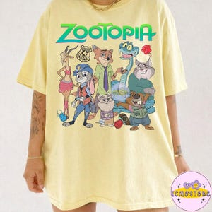 Pu&ograve; includere: T-shirt giallo chiaro con la scritta "ZOOTOPIA" in verde e blu, e personaggi dei cartoni animati del film. I personaggi includono un coniglio, una volpe, un serpente e altri. La maglietta ha un girocollo e maniche corte.