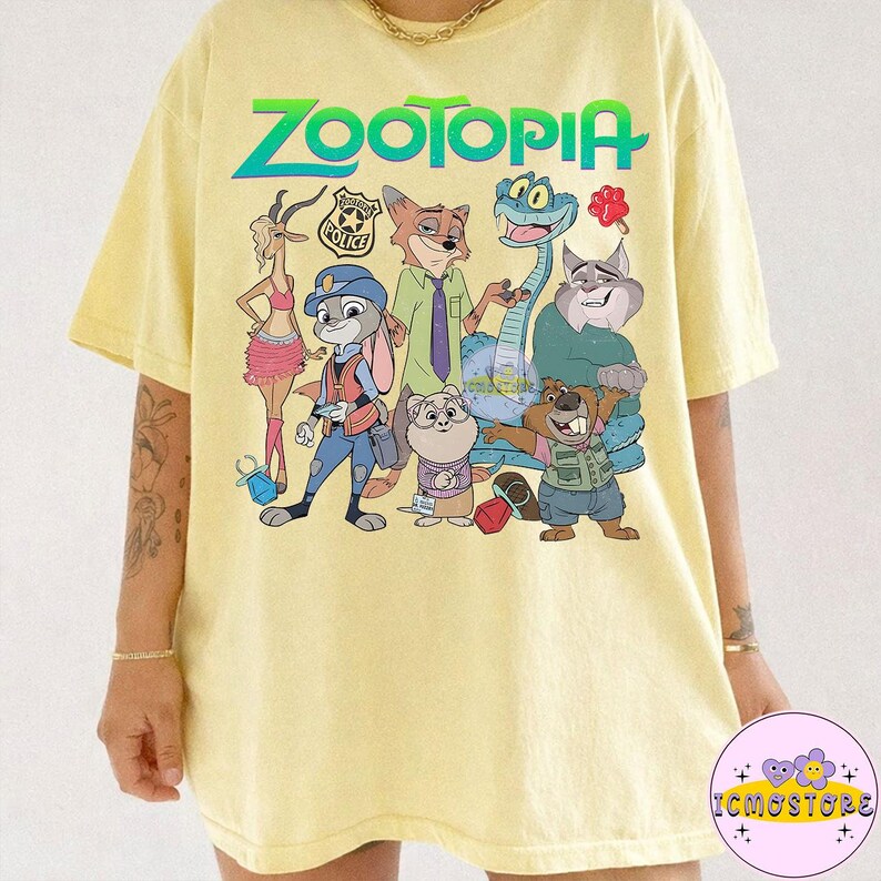 Pu&ograve; includere: T-shirt giallo chiaro con la scritta "ZOOTOPIA" in verde e blu, e personaggi dei cartoni animati del film. I personaggi includono un coniglio, una volpe, un serpente e altri. La maglietta ha un girocollo e maniche corte.