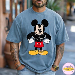 May include: A blue t-shirt featuring a muscular Mickey Mouse graphic. The cartoon character is depicted with black muscles, red shorts, yellow shoes, and white gloves. The shirt has short sleeves and a crew neck.