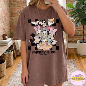 May include: Brown t-shirt featuring a group of Disney characters, including Mickey Mouse, Minnie Mouse, Donald Duck, and Goofy, against a black and white checkered background. The text "MICKEY & CO. EST 1928" is printed below the characters.