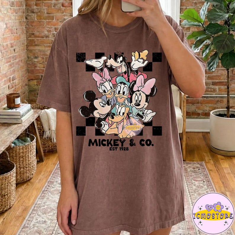May include: Brown t-shirt featuring a group of Disney characters, including Mickey Mouse, Minnie Mouse, Donald Duck, and Goofy, against a black and white checkered background. The text "MICKEY & CO. EST 1928" is printed below the characters.