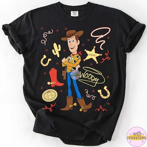 May include: Black t-shirt featuring a cartoon illustration of Woody from Toy Story. The design includes Woody in his cowboy attire, a sheriff's badge, a lasso, a cactus, a horseshoe, and the word "Woody".