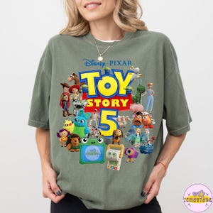 May include: Sage green t-shirt featuring a colorful graphic of characters from the Disney Pixar movie Toy Story 5. The design includes the movie title and the number 5 in yellow and red. The shirt is a relaxed fit.