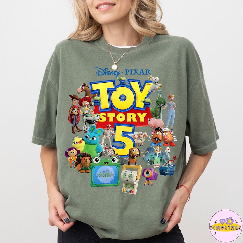 May include: Sage green t-shirt featuring a colorful graphic of characters from the Disney Pixar movie Toy Story 5. The design includes the movie title and the number 5 in yellow and red. The shirt is a relaxed fit.