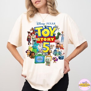 May include: Cream-colored t-shirt featuring a colorful graphic of characters from the Disney Pixar movie Toy Story 5. The design includes the movie title and the number 5, with various toy characters.