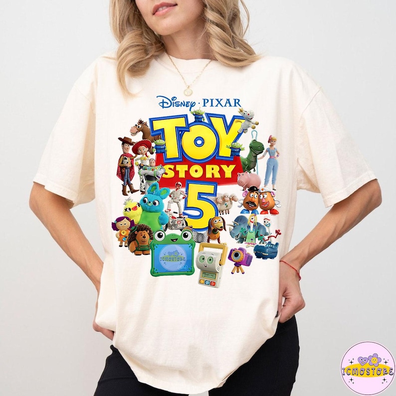 May include: Cream-colored t-shirt featuring a colorful graphic of characters from the Disney Pixar movie Toy Story 5. The design includes the movie title and the number 5, with various toy characters.