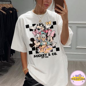 May include: White t-shirt featuring a graphic of Mickey Mouse and friends, including Minnie Mouse, Donald Duck, and Goofy. The design is set against a black and white checkered background, with the text "Mickey & Co. EST 1928".