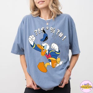 May include: A light blue t-shirt featuring a cartoon image of Donald Duck in a fit of anger, with symbols above his head. Donald Duck is wearing a blue sailor suit and a red bow tie. The shirt is a comfortable, oversized fit.