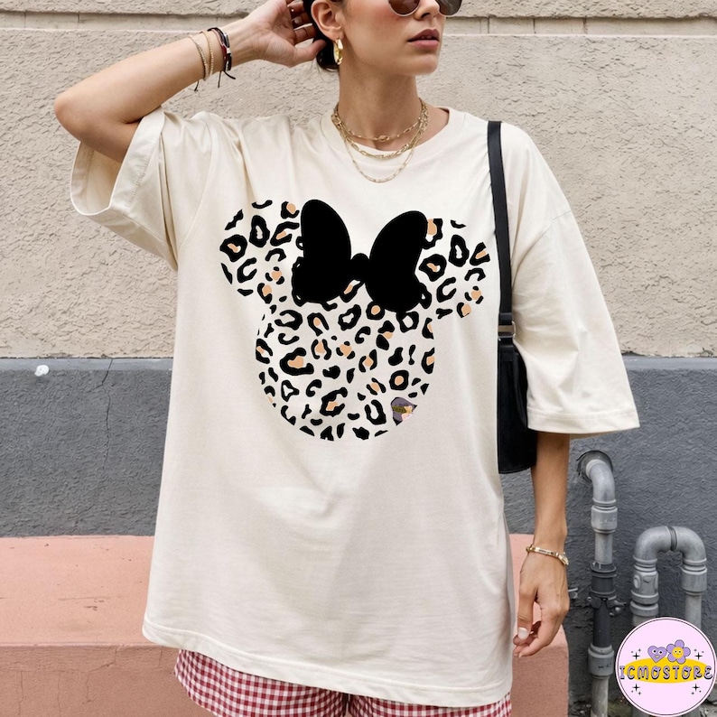 May include: Cream-colored t-shirt featuring a Minnie Mouse design with a leopard print head and a black bow. The shirt has short sleeves and a relaxed fit. The design is centered on the front of the shirt.