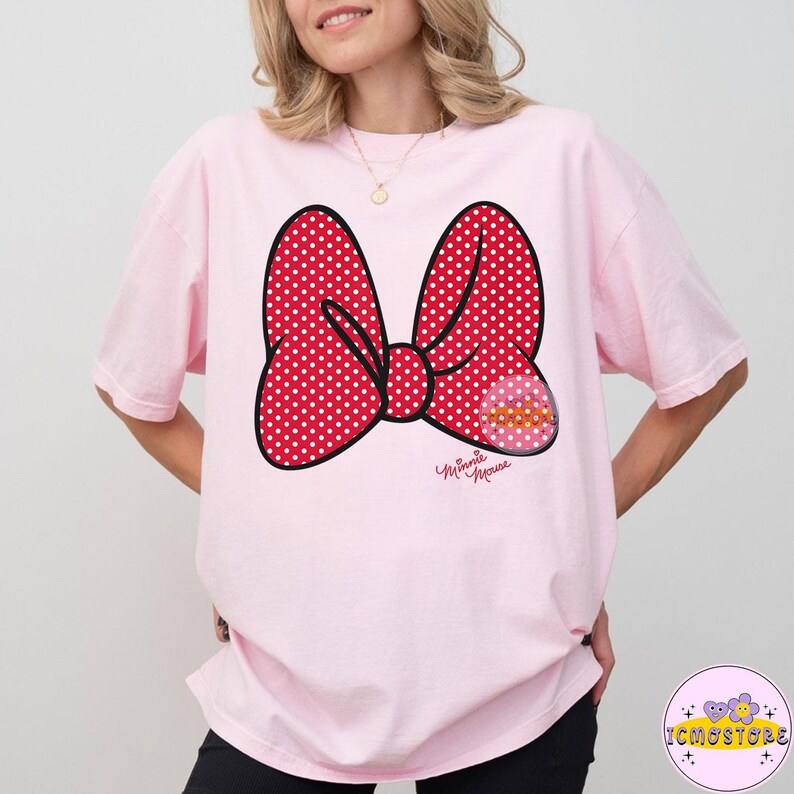 May include: A light pink t-shirt featuring a large red bow with white polka dots. The bow has a black outline and a small graphic with the words "Minnie Mouse" in cursive. The shirt is a casual style.