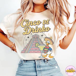 Puede incluir: Camiseta color crema con el texto "Cinco De Drinko" en escritura amarilla. El gráfico presenta un pato de dibujos animados con sombrero sosteniendo una margarita frente a una pirámide y una luna llena.